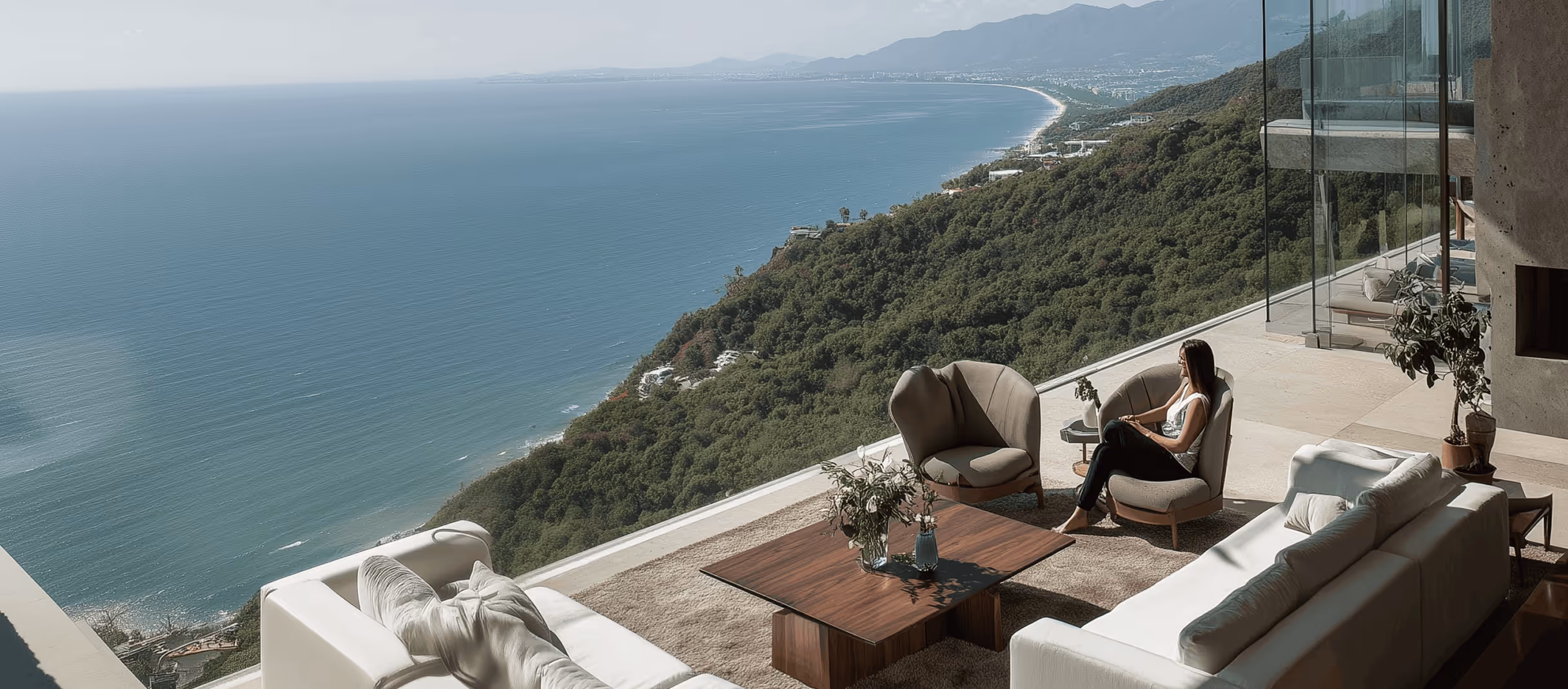 Modern terrace living area with white sofas, two armchairs, wooden table, and a woman sitting looking out over a scenic coastline and ocean.